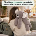 Dziecko siedzi bokiem na kanapie, łapki szarej koali przewieszone przez jego barki, obok plecak, twarz poza kadrem